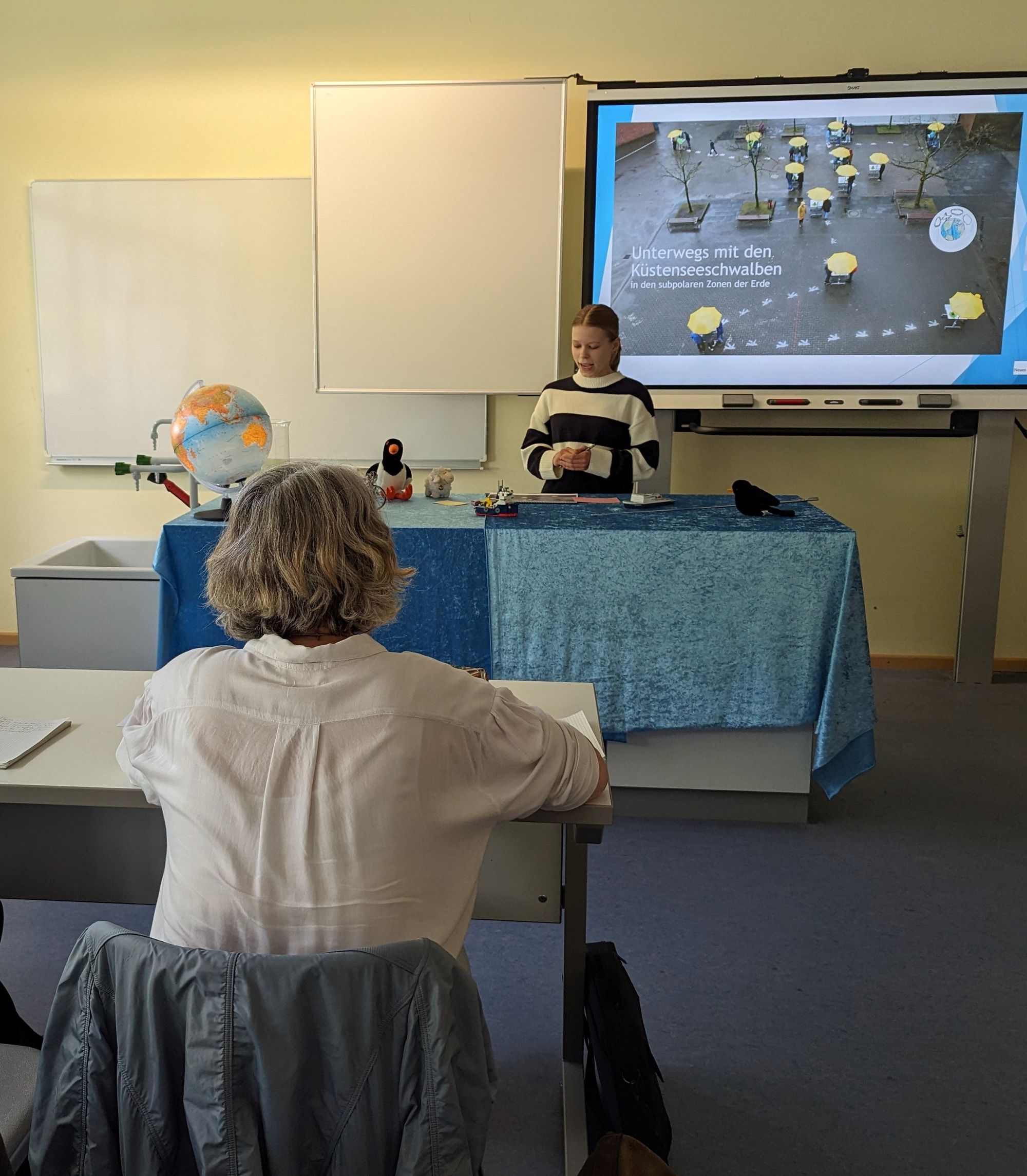 Susanne Menge von hinten, an einem Schultisch sitzend; vorne: eine Schülerin hält eine Präsentation