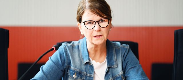 Susanne Menge sitzt vor einem Mikrofon mit ernstem Gesichtsausdruck; im Hintergrund ist verschwommen eine rote Wand zu sehen